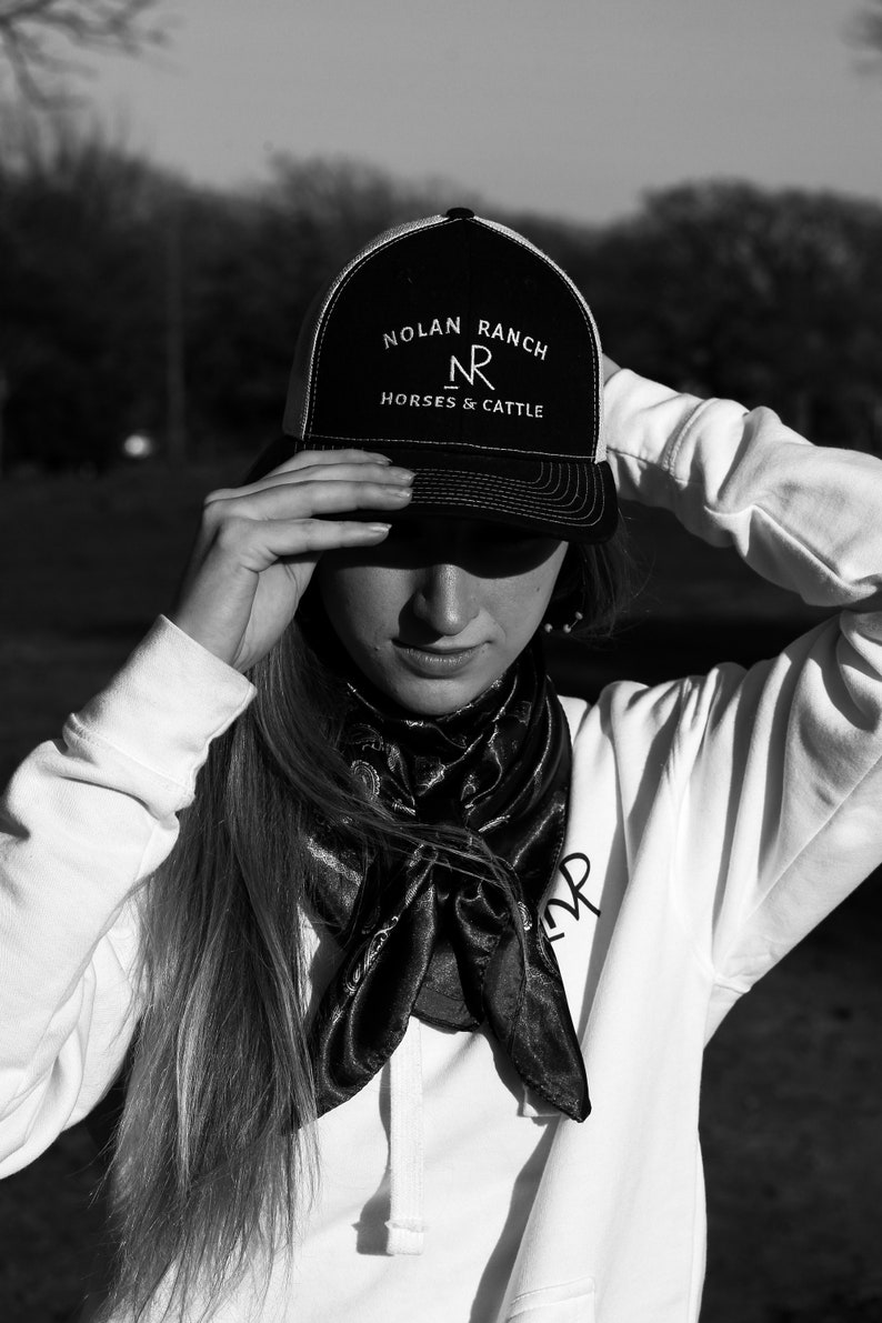 Może przedstawiać: Osoba w czarno-białej czapce trucker z napisem "Nolan Ranch" i "Horses & Cattle" z przodu. Osoba nosi r&oacute;wnież białą bluzę z kapturem i czarną chustkę.