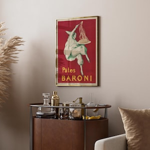 Op de afbeelding: Een ingelijste rechthoekige wandposter met een dieprode achtergrond en een witte illustratie van een figuur met een puntmuts die een bord pasta vasthoudt. De tekst P&acirc;tes BARONI staat in het geel onderaan de poster in een gouden lijst.