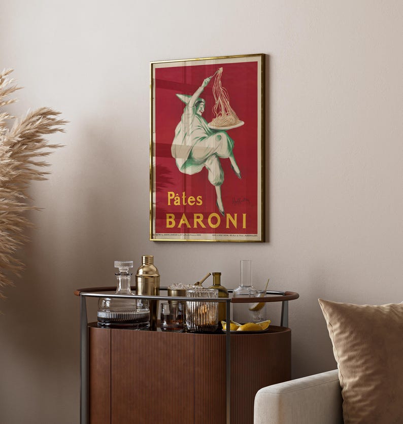 Op de afbeelding: Een ingelijste rechthoekige wandposter met een dieprode achtergrond en een witte illustratie van een figuur met een puntmuts die een bord pasta vasthoudt. De tekst P&acirc;tes BARONI staat in het geel onderaan de poster in een gouden lijst.