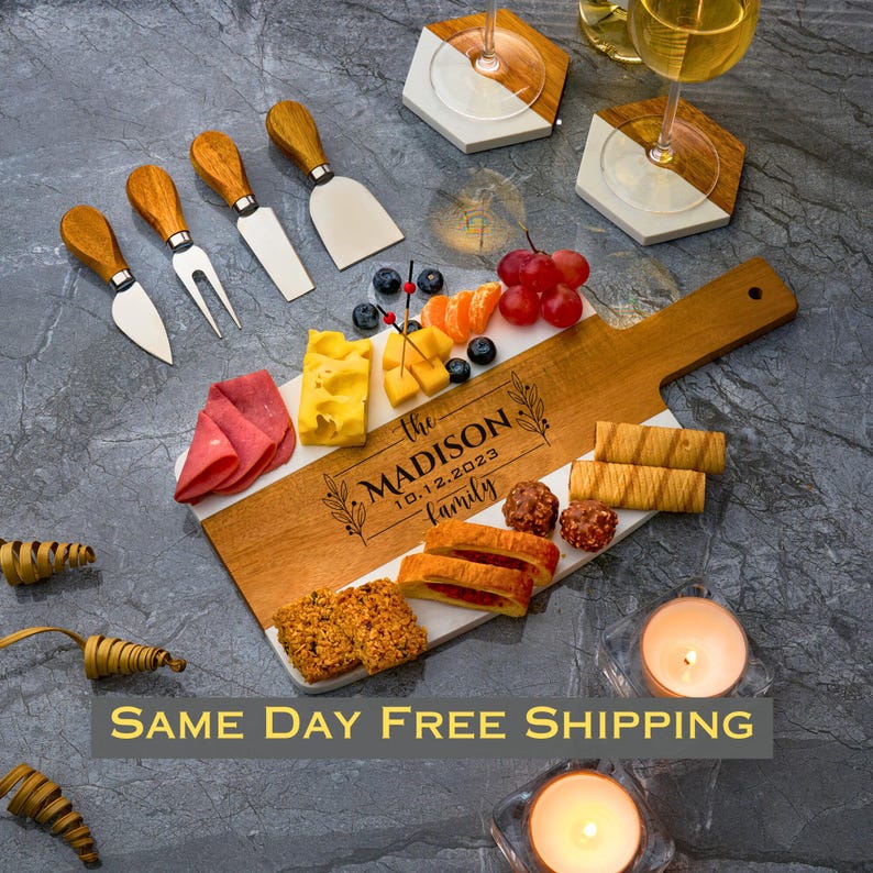 May include: A white and wood serving board with a handle. The board has a white marble-like surface with a wood border. The board is decorated with the text "The Madison Family 10.12.2023". The board is surrounded by various snacks, including cheese, crackers, fruit, and chocolate.