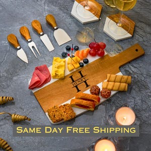 May include: Personalized wooden and marble cheese board with a handle, featuring the name "Henry" engraved in a double-H monogram design. The board is filled with various cheeses, fruits, crackers, and other snacks.  The board is accompanied by a set of five cheese knives with wooden handles.