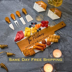 May include: A white and brown rectangular serving board with a wooden handle. The board has a personalized inscription that reads "The Fletchers 40°23′17″ N 74°19′42". The board is filled with various snacks, including cheese, crackers, fruit, and chocolate.
