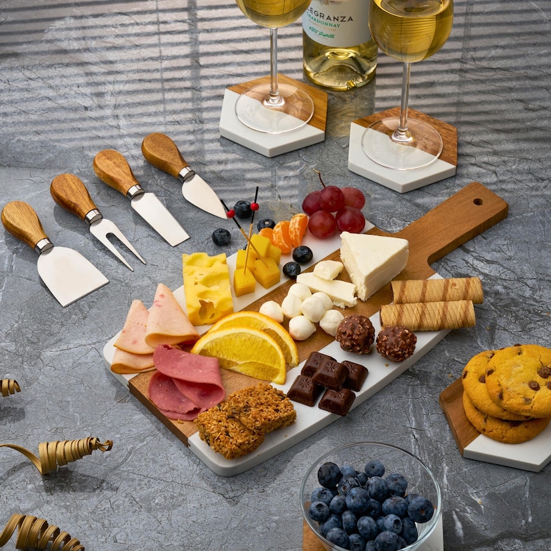 May include: A white and wood serving board with a variety of snacks, including cheese, fruit, crackers, and chocolate. The board is surrounded by two wine glasses and coasters.