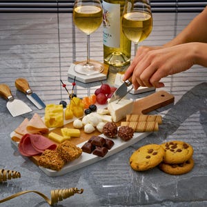 May include: A white marble cheese board with a variety of cheeses, crackers, grapes, and chocolate. The board is on a gray countertop with two glasses of white wine and a bottle of white wine.