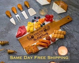 May include: A rectangular wooden and white serving board engraved with "Shana Tova" and "The Katz Family" featuring cheese, fruit, and crackers. Includes four cheese knives with wooden handles, two hexagonal coasters, and two tea light candles.