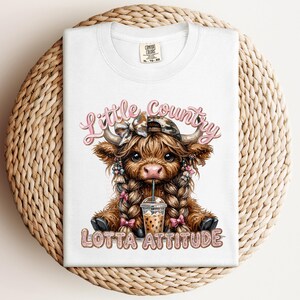 May include: White Comfort Colors t-shirt featuring a cartoon Highland cow with braids, a bow, and a drink. The text "Little Country Lotta Attitude" is displayed in pink and glittery lettering. The shirt is folded on a woven surface.