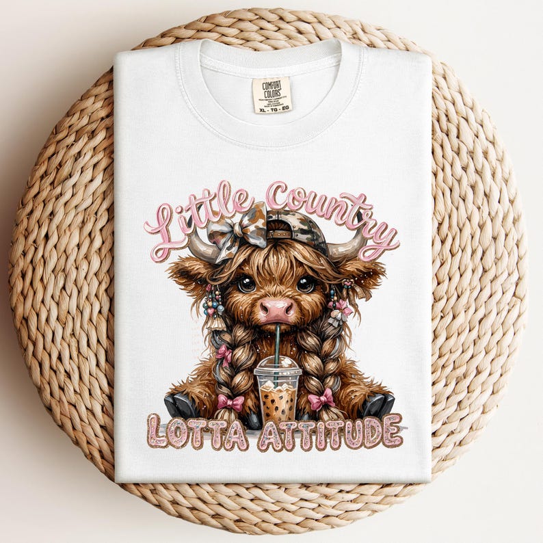 May include: White Comfort Colors t-shirt featuring a cartoon Highland cow with braids, a bow, and a drink. The text "Little Country Lotta Attitude" is displayed in pink and glittery lettering. The shirt is folded on a woven surface.