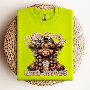 May include: A neon green sweatshirt featuring a graphic of a Highland cow with braids, a camo hat, and a drink. The text reads "Little Country Lotta Attitude" in pink script. The sweatshirt is folded on a woven mat.