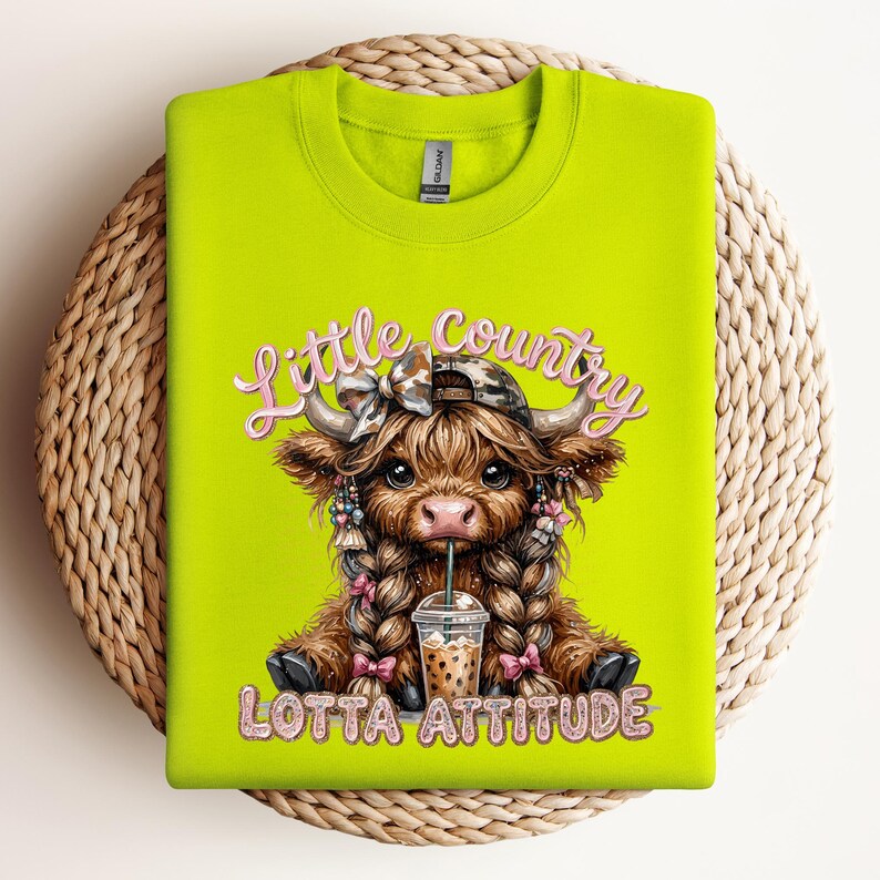 May include: A neon green sweatshirt featuring a graphic of a Highland cow with braids, a camo hat, and a drink. The text reads "Little Country Lotta Attitude" in pink script. The sweatshirt is folded on a woven mat.