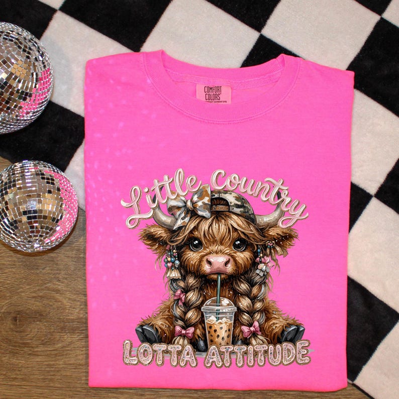 May include: A hot pink Comfort Colors t-shirt featuring a cartoon Highland cow with a camo hat, braids, and a drink. The text "Little Country" and "Lotta Attitude" are in a glittery font. The shirt is on a black and white checkered surface.