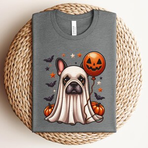 Pu&ograve; includere: Una maglietta grigia con una simpatica illustrazione di un bulldog francese vestito da fantasma per Halloween. Il bulldog indossa un costume da fantasma bianco con un palloncino a forma di zucca e pipistrelli e stelle sullo sfondo.