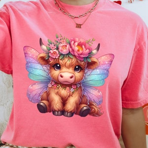 K&ouml;nnte beinhalten: Ein pinkfarbenes T-Shirt mit einem Cartoon-Bild einer braunen Highland-Kuh mit Feenfl&uuml;geln. Die Kuh tr&auml;gt eine Blumenkrone und eine Blumenkette. Die Fl&uuml;gel sind schillernd und haben einen Regenbogenfarbton.