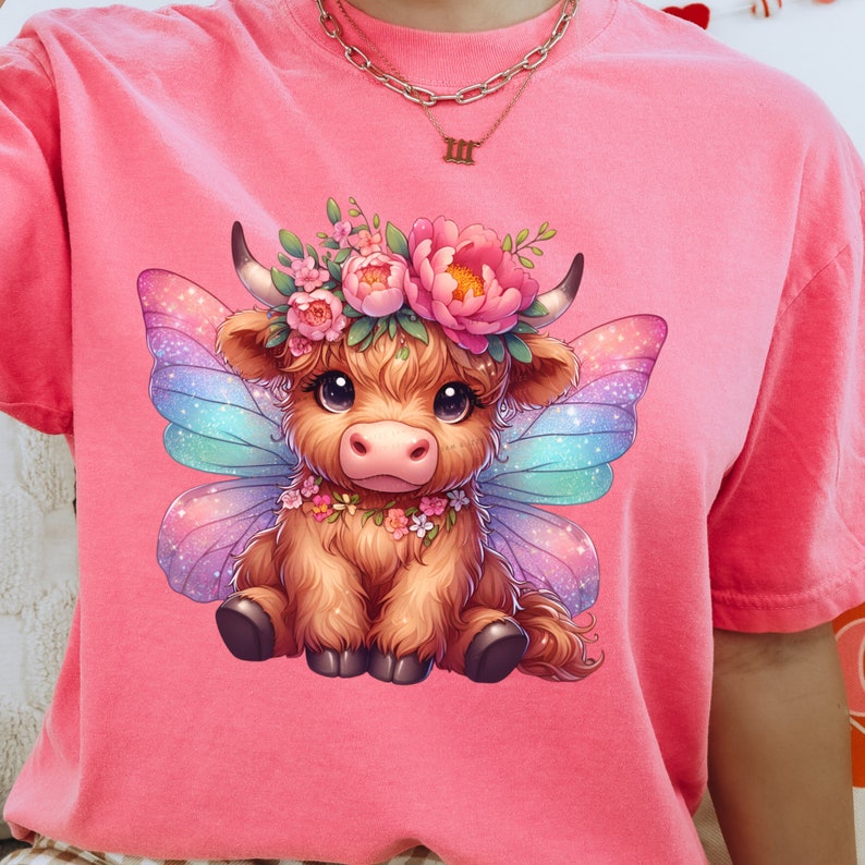 K&ouml;nnte beinhalten: Ein pinkfarbenes T-Shirt mit einem Cartoon-Bild einer braunen Highland-Kuh mit Feenfl&uuml;geln. Die Kuh tr&auml;gt eine Blumenkrone und eine Blumenkette. Die Fl&uuml;gel sind schillernd und haben einen Regenbogenfarbton.