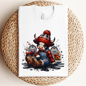 May include: White t-shirt featuring a cartoon illustration of a cow wearing a red and black plaid hat, overalls, and boots. The cow is sitting in front of a barn and tractor. The shirt is folded on a woven surface.