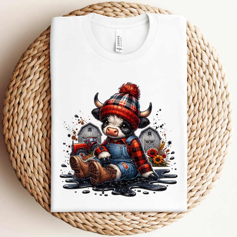 May include: White t-shirt featuring a cartoon illustration of a cow wearing a red and black plaid hat, overalls, and boots. The cow is sitting in front of a barn and tractor. The shirt is folded on a woven surface.