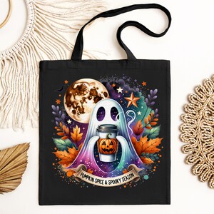 K&ouml;nnte beinhalten: Eine schwarze Tragetasche mit einer farbenfrohen Illustration eines Geistes, der einen K&uuml;rbisgew&uuml;rz-Latte h&auml;lt. Der Geist ist umgeben von Sternen, einem Halbmond und Herbstbl&auml;ttern. Der Text "Pumpkin Spice & Spooky Season" ist auf einem Banner unter dem Geist gedruckt.