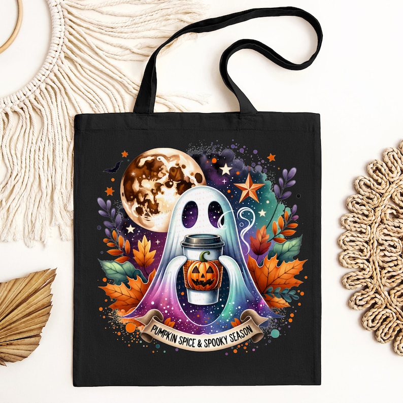 K&ouml;nnte beinhalten: Eine schwarze Tragetasche mit einer farbenfrohen Illustration eines Geistes, der einen K&uuml;rbisgew&uuml;rz-Latte h&auml;lt. Der Geist ist umgeben von Sternen, einem Halbmond und Herbstbl&auml;ttern. Der Text "Pumpkin Spice & Spooky Season" ist auf einem Banner unter dem Geist gedruckt.