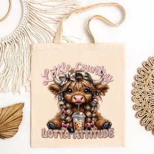 May include: Beige tote bag featuring a cartoon Highland cow with braids, a camo hat, and a drink. The text "Little Country Lotta Attitude" is displayed in pink and glittery letters. The bag has a simple design with two handles.