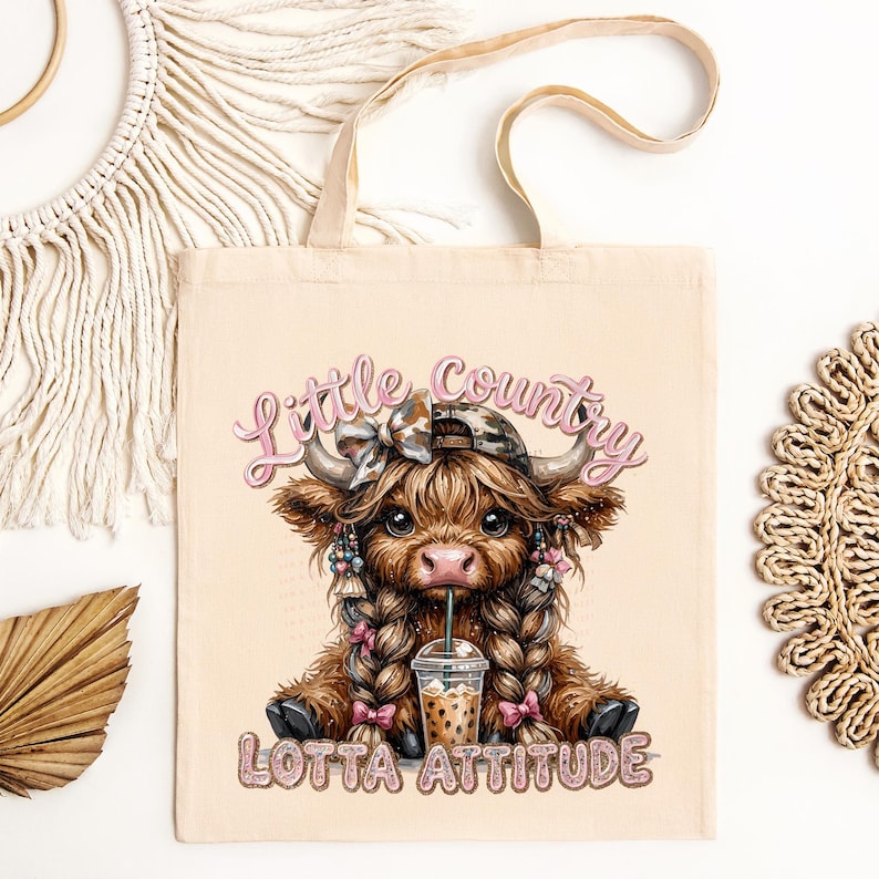 May include: Beige tote bag featuring a cartoon Highland cow with braids, a camo hat, and a drink. The text "Little Country Lotta Attitude" is displayed in pink and glittery letters. The bag has a simple design with two handles.