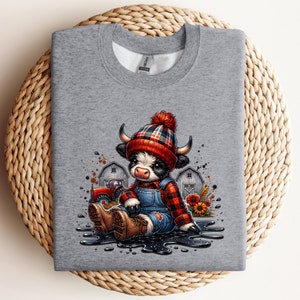 May include: A heather gray sweatshirt featuring a cartoon illustration of a cow wearing a plaid hat and overalls. The cow is sitting in a puddle with a tractor and barns in the background. The design has a farm theme.