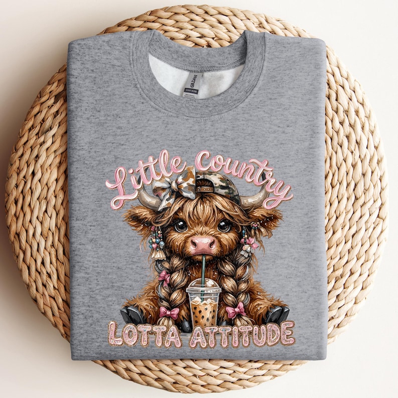 May include: A heather gray sweatshirt featuring a graphic of a Highland cow with braids, a camo hat, and a drink. The text "Little Country Lotta Attitude" is in pink script. The sweatshirt is on a woven basket.