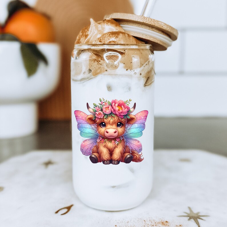 K&ouml;nnte beinhalten: Ein durchsichtiger Glasbecher mit einem braunen und wei&szlig;en, schaumigen Getr&auml;nk. Der Becher hat eine niedliche Cartoon-Illustration einer braunen Kuh mit rosa und blauen Schmetterlingsfl&uuml;geln und einer Blumenkrone.