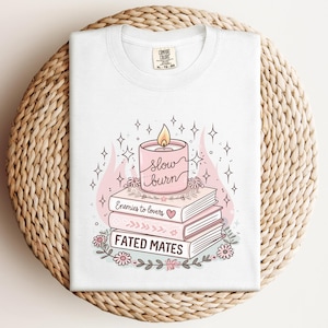May include: White t-shirt featuring a graphic of a pink candle with the words "slow burn" above a stack of books. The books are titled "Enemies to lovers" and "Fated mates". Floral and star accents complete the design.