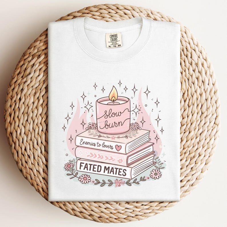 May include: White t-shirt featuring a graphic of a pink candle with the words "slow burn" above a stack of books. The books are titled "Enemies to lovers" and "Fated mates". Floral and star accents complete the design.