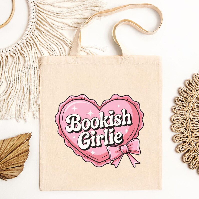 Op de afbeelding: Een roze canvas draagtas met een roze hartontwerp en de tekst "Bookish Girlie" in wit. Het hart heeft een witte strik en glitters.