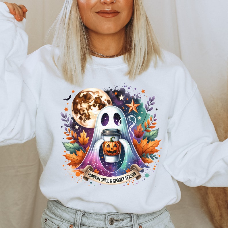 K&ouml;nnte beinhalten: Wei&szlig;er Pullover mit einem Aufdruck eines Geistes, der einen K&uuml;rbisgew&uuml;rz-Latte h&auml;lt. Der Aufdruck enth&auml;lt auch einen Vollmond, Sterne und Herbstbl&auml;tter. Der Text "Pumpkin Spice & Spooky Season" ist unter dem Aufdruck gedruckt.