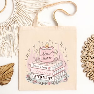 May include: Cream canvas tote bag with a pink candle design, the words "slow burn", and a flame. Below the candle are three books with the titles "Enemies to lovers" and "Fated mates". The design includes floral and star accents.