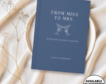 Op de afbeelding: Een marineblauw notitieboek met de tekst "FROM MISS TO MRS." en "NOTES FOR THE NEXT CHAPTER." Het notitieboek bevat een afbeelding van twee champagneglazen en de naam "OLIVIA ANDERSON." De afbeelding bevat ook een wit bord en decoratieve takjes.