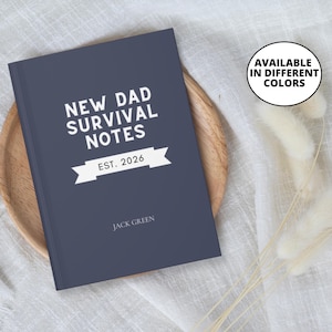 Puede incluir: Un cuaderno azul marino con el texto "NEW DAD SURVIVAL NOTES" en blanco. Debajo del título está "EST. 2026" en una pancarta blanca. El nombre "JACK GREEN" está impreso en la parte inferior. El cuaderno está sobre una bandeja de madera.