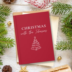 May include: A red Christmas card with the text "CHRISTMAS with the Johnsons" and a white Christmas tree illustration. The card is on top of a cream-colored envelope, with pine needles and ornaments in the background.