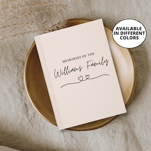 May include: A light beige family memory book with the text "Memories of the Williams Family" in elegant script. Two small hearts are drawn below the family name. The book is displayed on a gold-coloured plate.