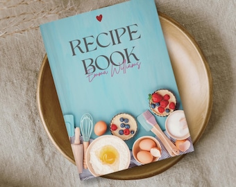 May include: A recipe book titled "Recipe Book Emma Williams" with a teal cover and a red heart. The book is on a gold plate with baking ingredients and pastries. A whisk, eggs, and a rolling pin are visible.