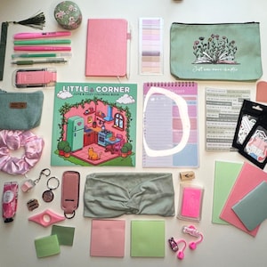 May include: A collection of stationery and accessories, including a pink notebook, pens, a coloring book, a planner, and a headband. The items are in shades of pink, green, and neutral tones, arranged on a white surface.