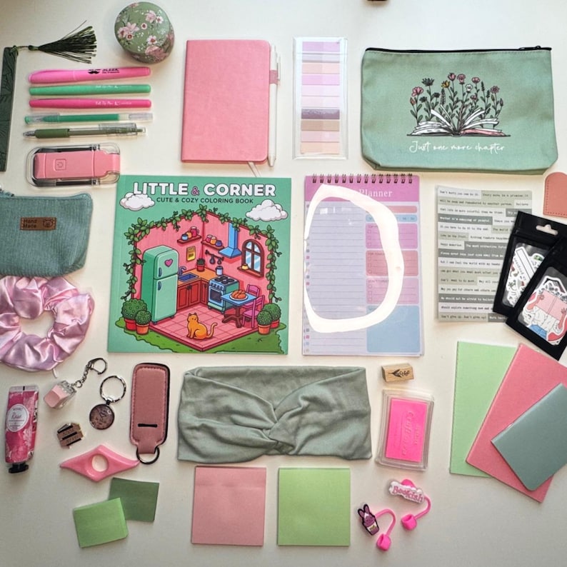 May include: A collection of stationery and accessories, including a pink notebook, pens, a coloring book, a planner, and a headband. The items are in shades of pink, green, and neutral tones, arranged on a white surface.