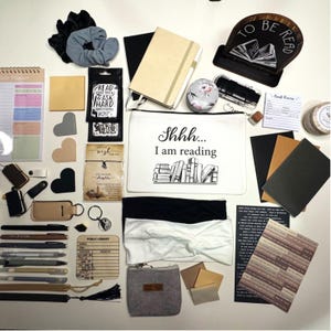 May include: A collection of reading and stationery items, including notebooks, pens, bookmarks, and a zippered pouch with the text "Shhh... I am reading." Also visible are scrunchies, headbands, and a wooden book stand.