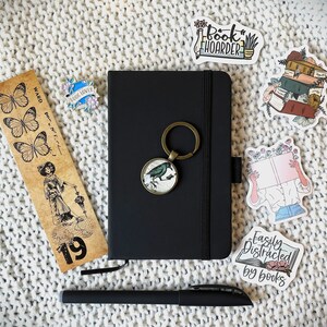 May include: A black notebook with a metal key chain attached to it. The notebook has a black cover and a white elastic band. The key chain has a small metal bird charm. The notebook is surrounded by various book-themed stickers, including "Book Hoarder", "Easily Distracted by Books", and a sticker of a person reading a book. There is also a bookmark with butterflies and the text "Ward, per. et. lit. - 19".