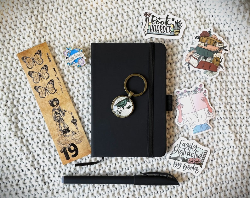 May include: A black notebook with a metal key chain attached to it. The notebook has a black cover and a white elastic band. The key chain has a small metal bird charm. The notebook is surrounded by various book-themed stickers, including "Book Hoarder", "Easily Distracted by Books", and a sticker of a person reading a book. There is also a bookmark with butterflies and the text "Ward, per. et. lit. - 19".
