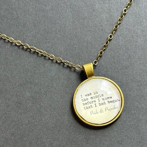 May include: A round pendant necklace with an antique brass finish featuring a white insert printed with the text: I was in the middle before I knew that I had begun. Pride & Prejudice. The pendant hangs from a matching brass-toned metal chain.