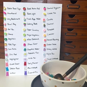 May include: A white sheet displays stationery items such as bookmarks and pens, each adorned with a decorative charm. A bowl with gold polka dots contains more charms and a pen. The background features wooden drawers.