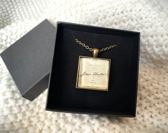 May include: A gold necklace with a square pendant featuring the title "Pride and Prejudice" and Jane Austen's signature in black ink.