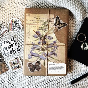 May include: A brown paper-wrapped journal with a floral design and a butterfly sticker. The journal is tied with twine and has a quote about developing knowledge. There are several stickers on the journal, including one that says "read more books".