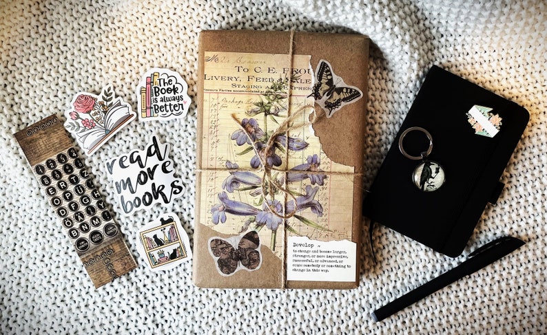 May include: A brown paper-wrapped journal with a floral design and a butterfly sticker. The journal is tied with twine and has a quote about developing knowledge. There are several stickers on the journal, including one that says "read more books".