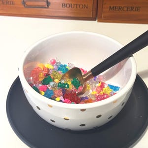 May include: A white ceramic bowl with gold polka dots filled with colourful beads. A gold-coloured scoop with a black handle rests inside the bowl. The bowl sits on a dark blue plate.