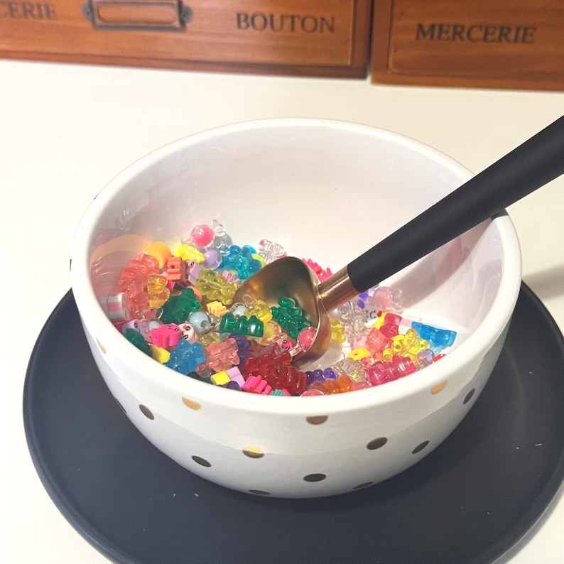 May include: A white ceramic bowl with gold polka dots filled with colourful beads. A gold-coloured scoop with a black handle rests inside the bowl. The bowl sits on a dark blue plate.