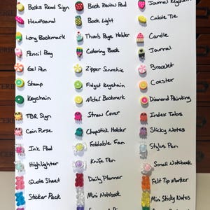 May include: A white board featuring a handwritten list of stationery items, each accompanied by a decorative charm. Items include a pencil bag, journal, keychain, and sticker pack. The charms are shaped like fruits, sweets, and other small objects.
