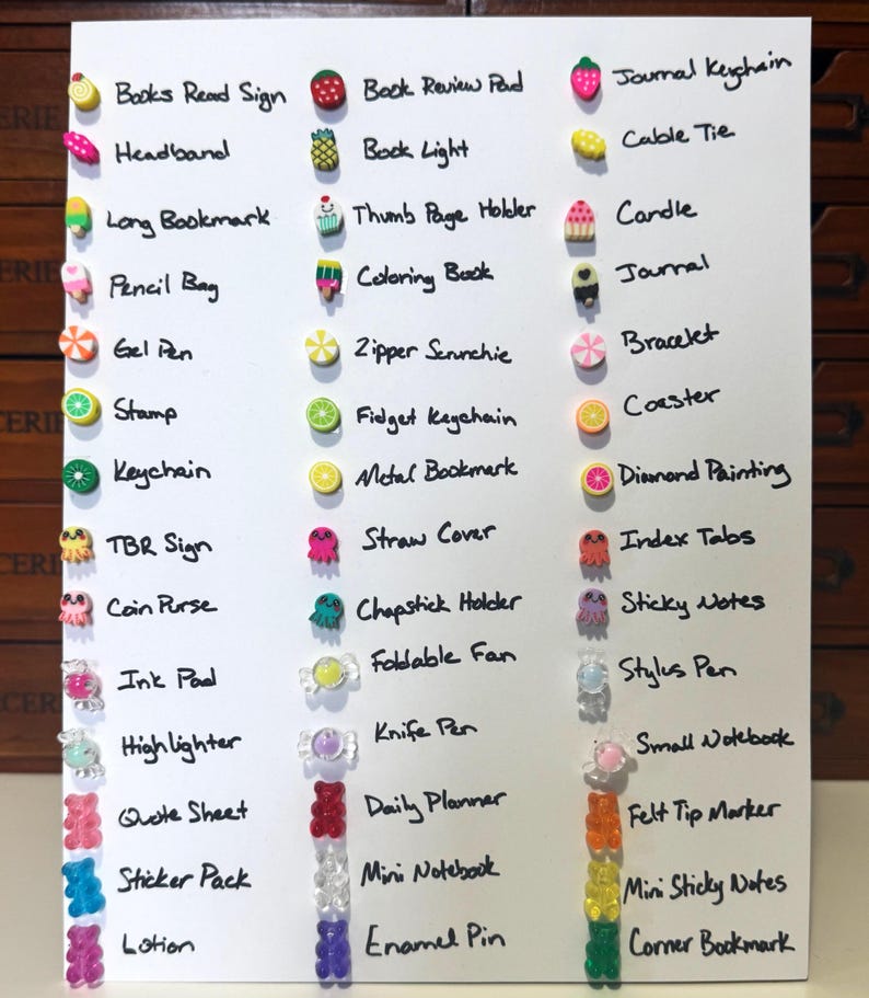 May include: A white board featuring a handwritten list of stationery items, each accompanied by a decorative charm. Items include a pencil bag, journal, keychain, and sticker pack. The charms are shaped like fruits, sweets, and other small objects.